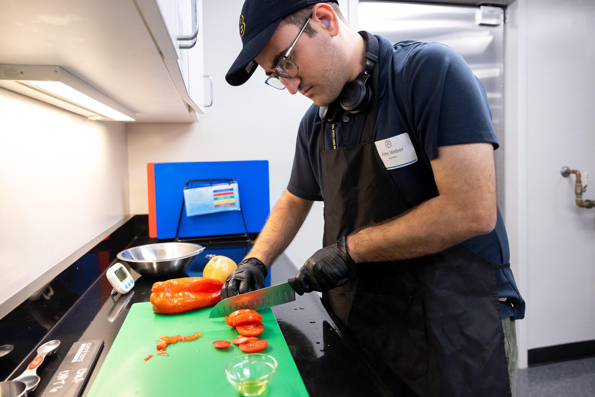 A Summer LIFE student wearing an apron and gloves slices a red bell pepper in a kitchen.