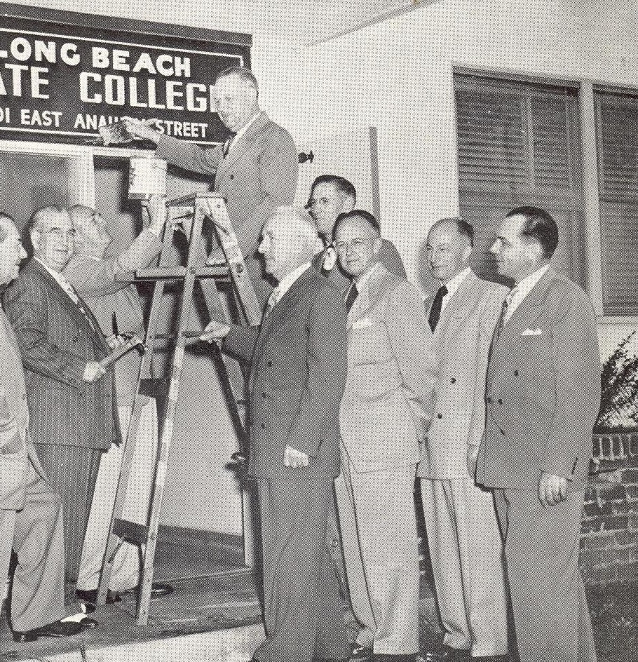 A group of men in suits gathers around a man on a ladder, holding a paintbrush near a sign that reads "Long Beach State College."