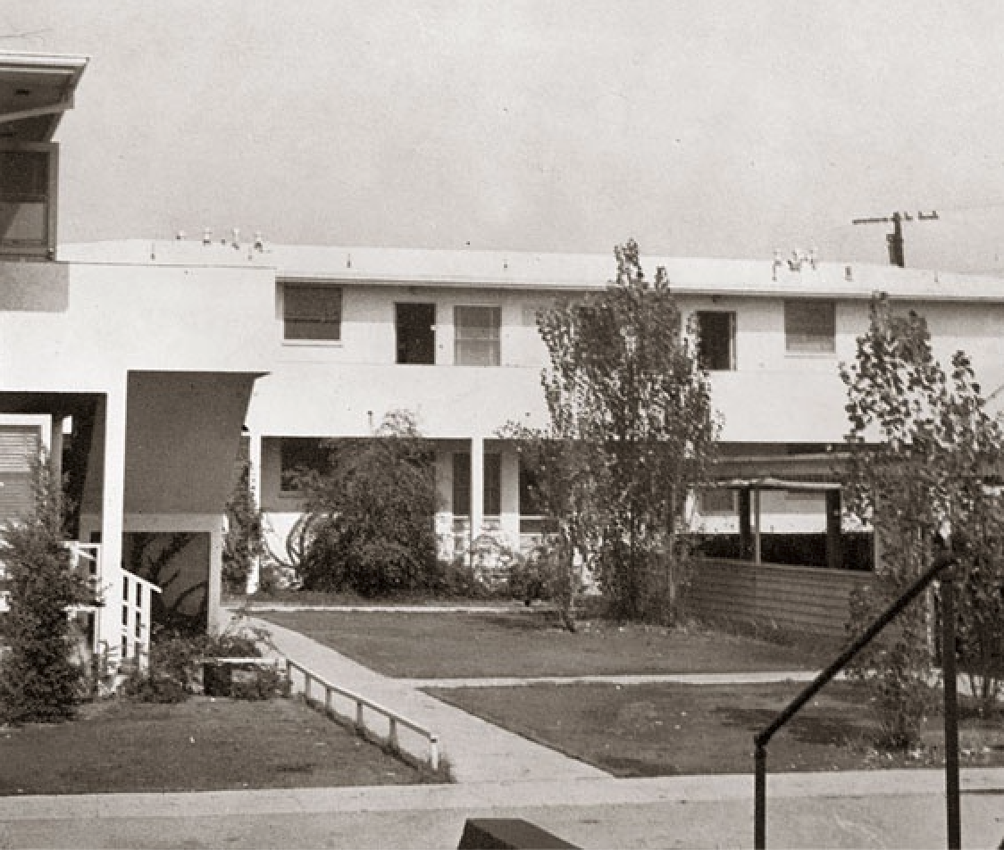 A two-story building with white walls and large windows stands in the background, with a small lawn and pathway leading toward it.