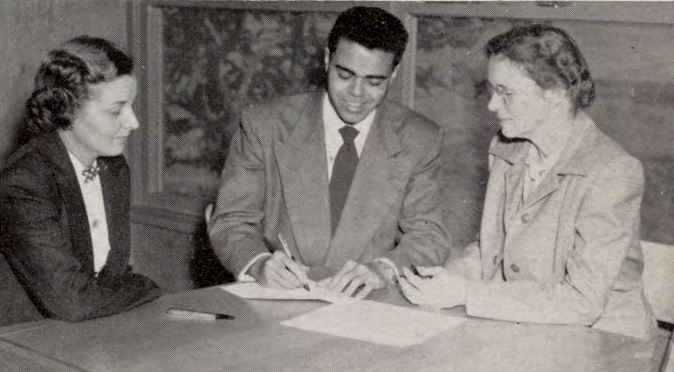 Roger Bryson signs a document at a table, flanked by two others, all dressed in business attire.