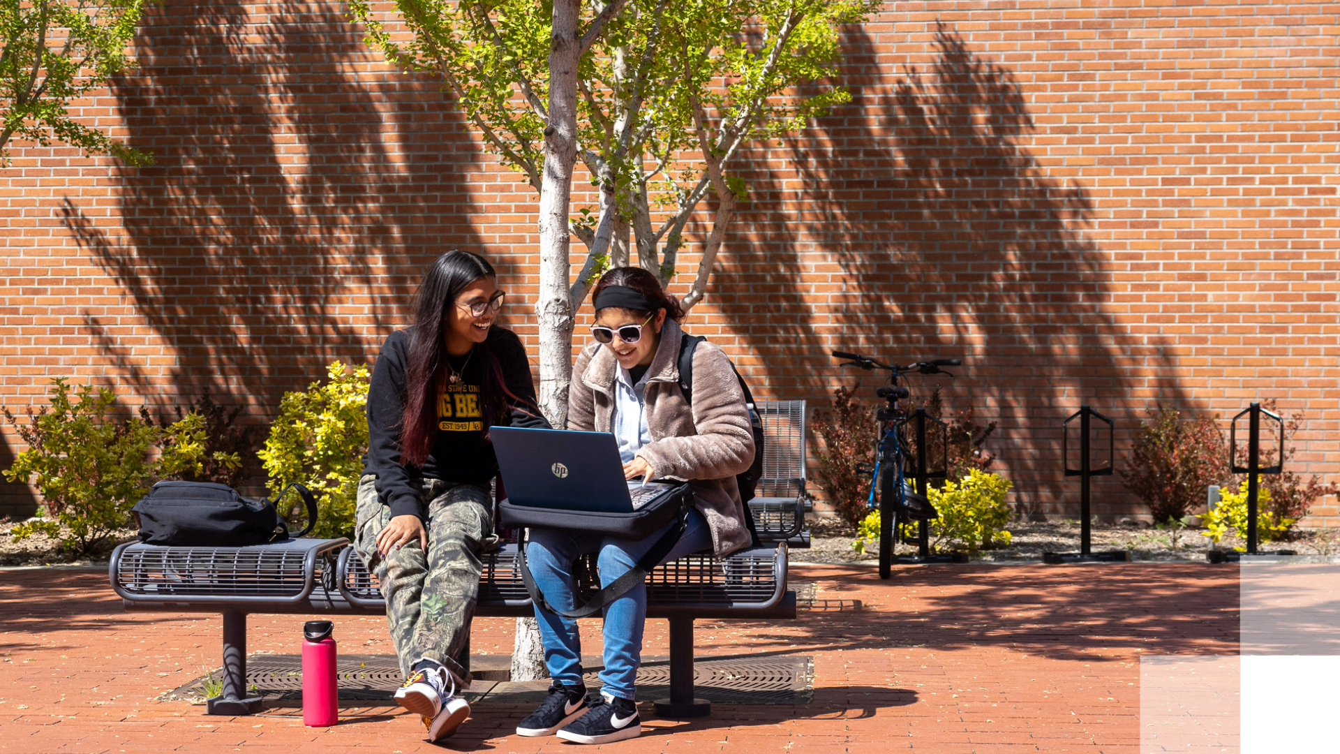 Two students sit on a bench outside and look at a laptop.