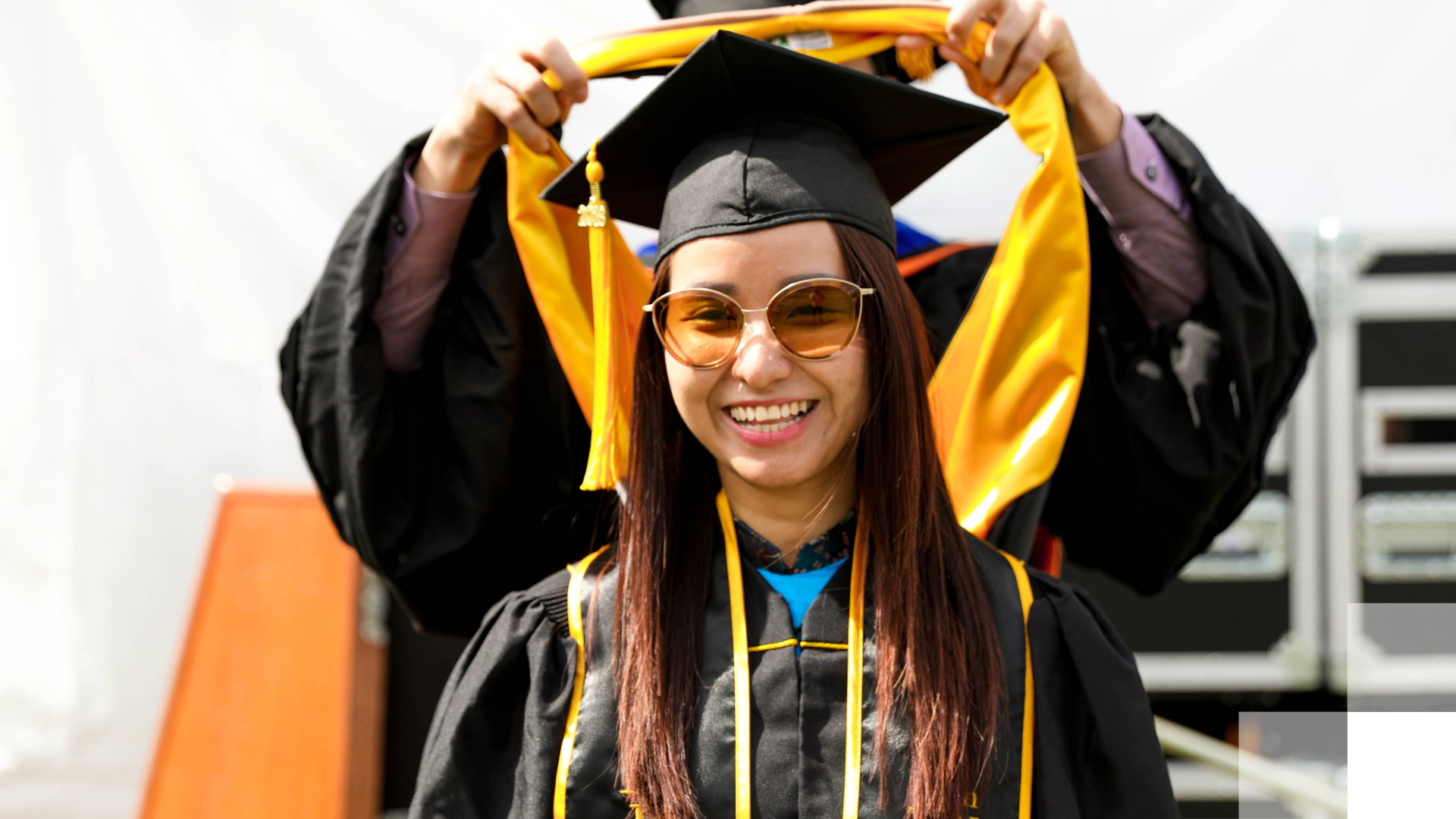 A graduate student in a black robe stands as someone behind her puts a graduate hood over her cap. 