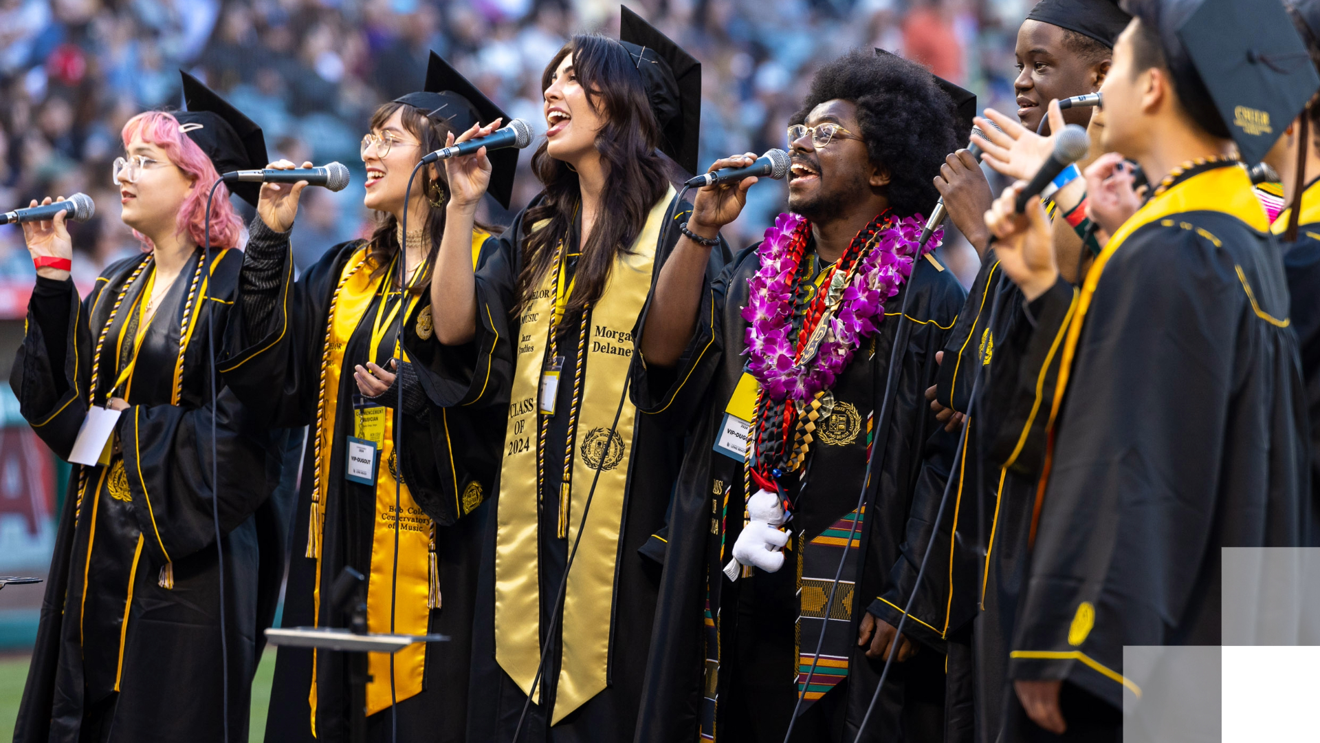 A group of students in caps and gowns sing to a crowd at graudation.