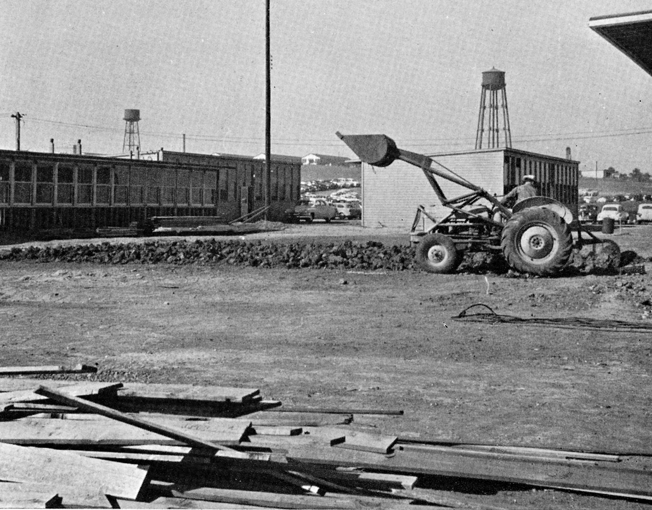 A tractor moves dirt near temporary buildings, with two water towers in the background.