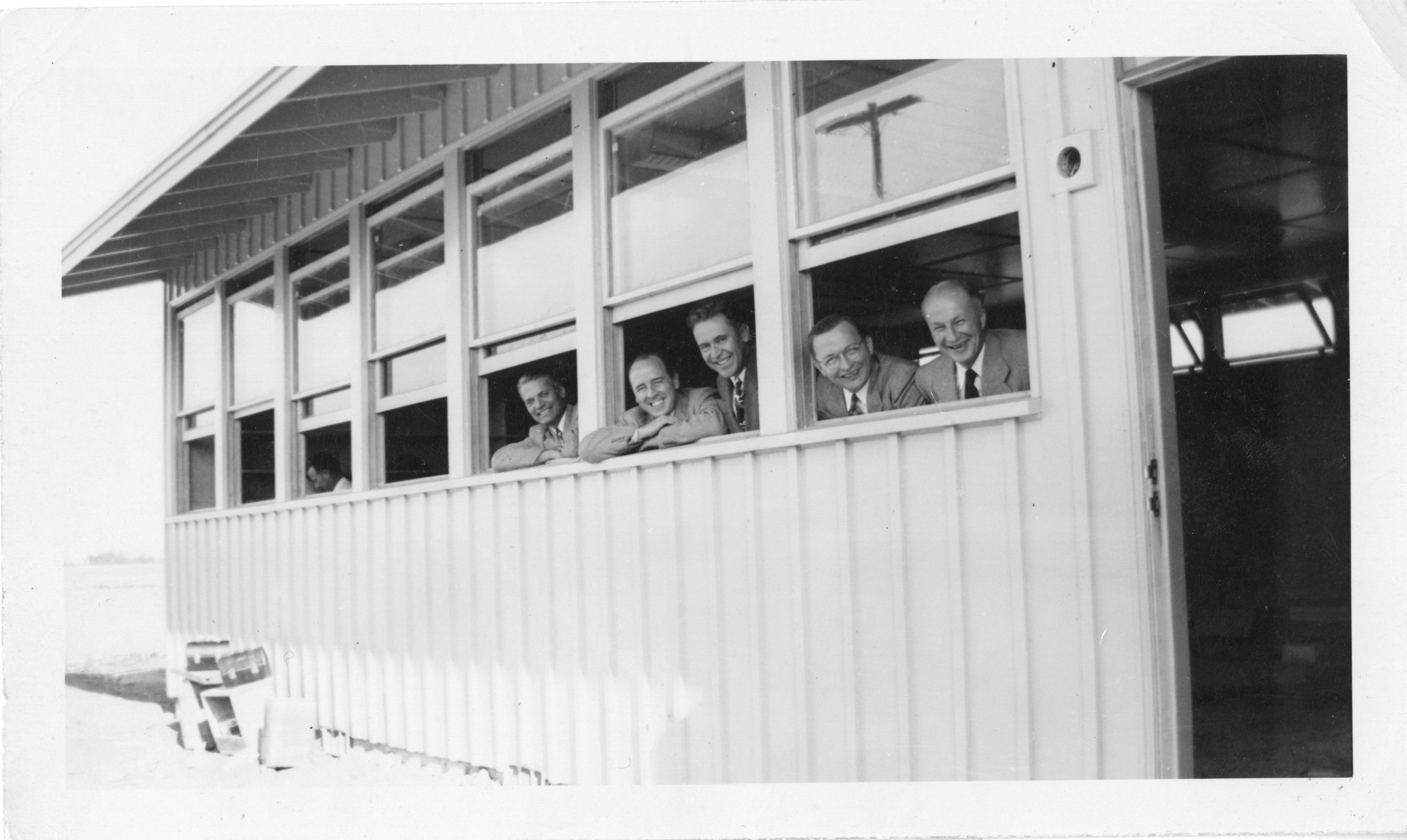 Five men in suits lean out of windows, smiling from a building with horizontal paneling.