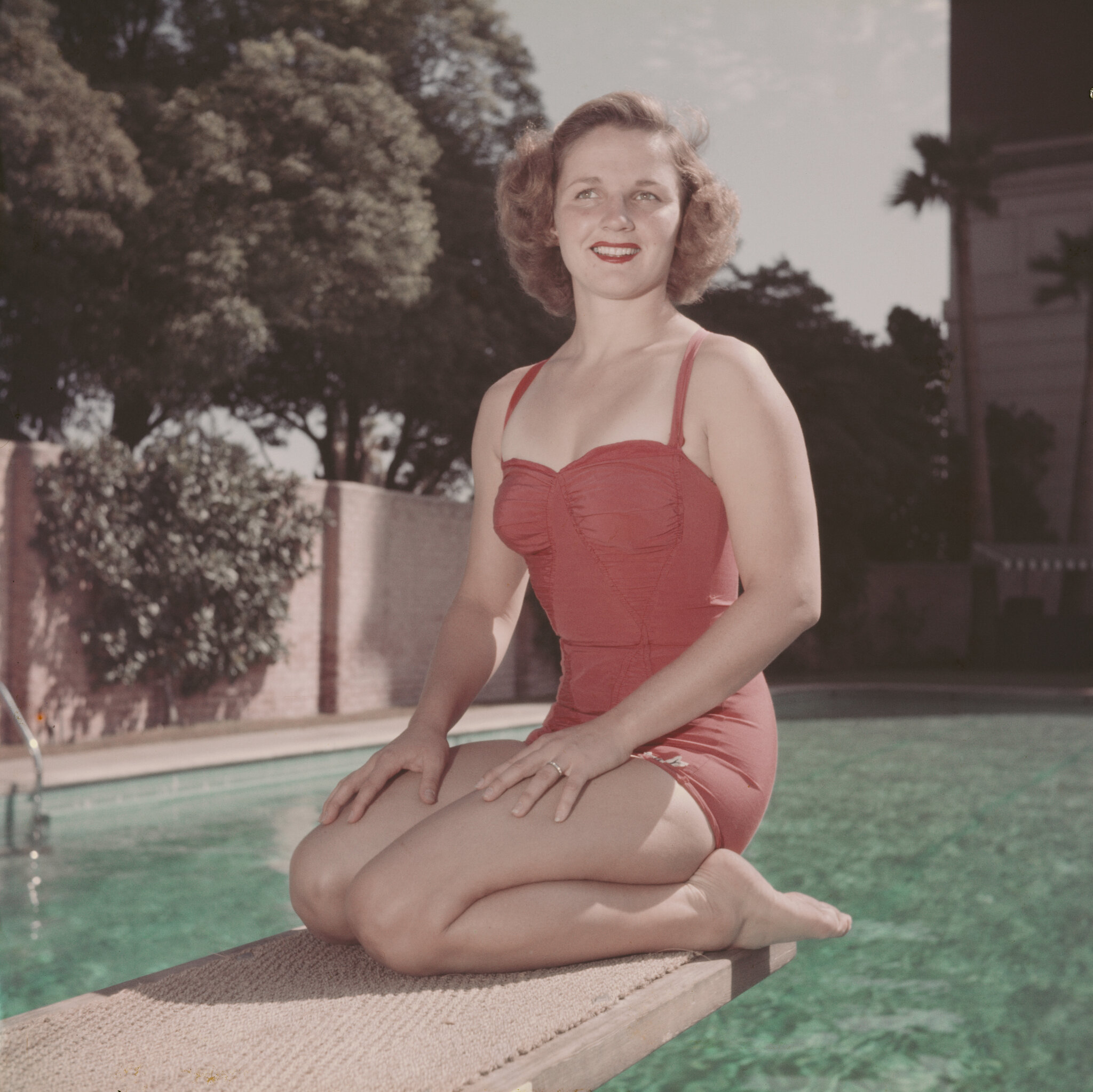 A person in a red swimsuit kneels on a diving board by a pool, smiling at the camera.