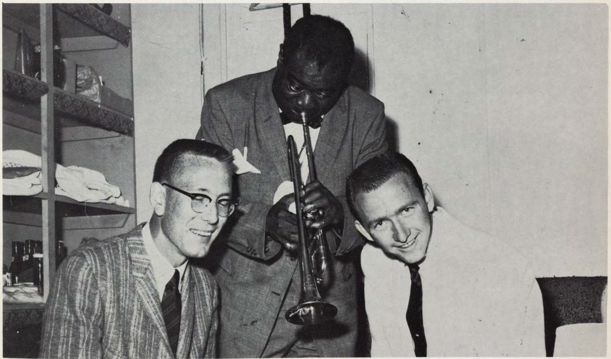 A trumpet player stands between two smiling men in a small room.