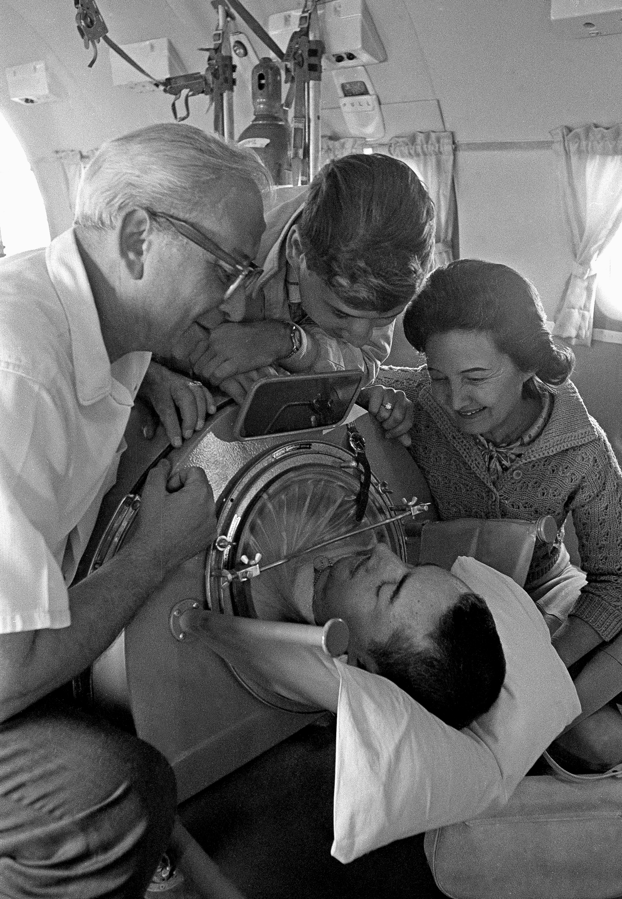 Three people smile and look at a person inside an iron lung, engaged in conversation.