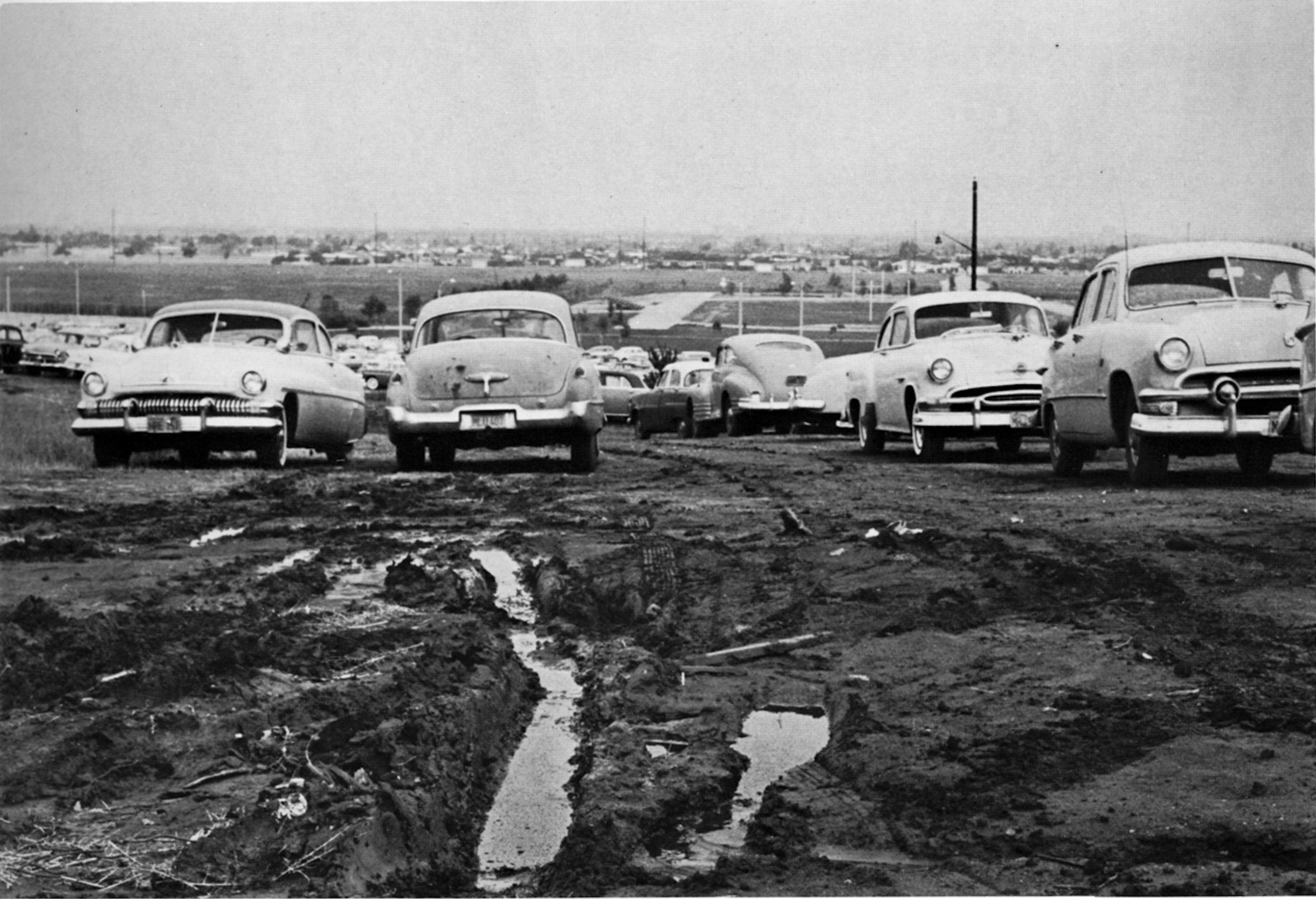 Cars are parked on a muddy dirt lot, with deep tire tracks visible in the foreground.