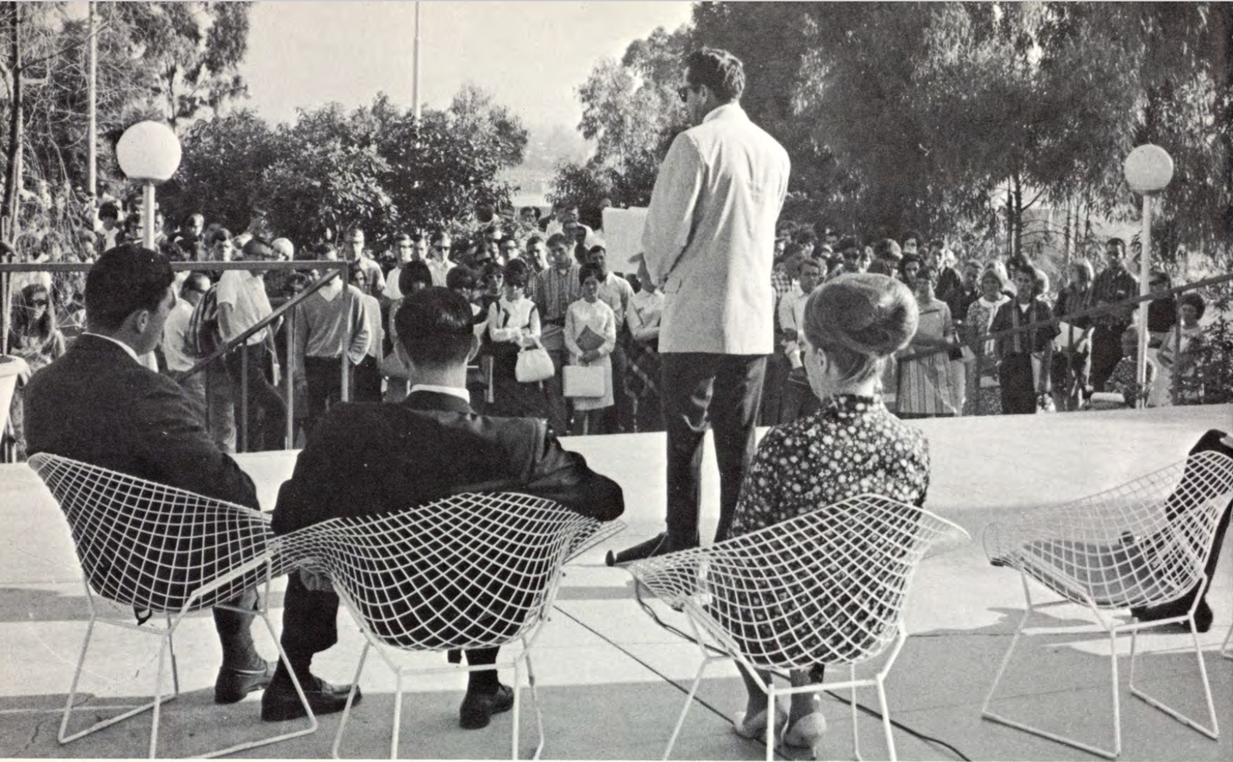 A speaker faces a large audience while three seated people, seated in white metal chairs, listen from behind.