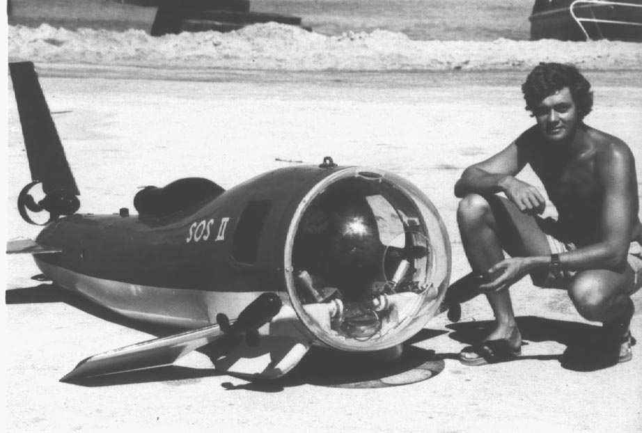 A person crouches next to a small personal submersible on the beach.