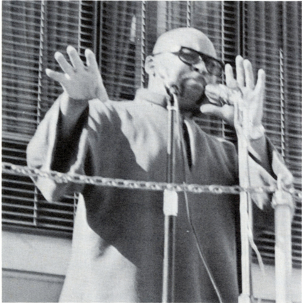 Maulana Karenga person with glasses speaks passionately, raising both hands at a microphone.