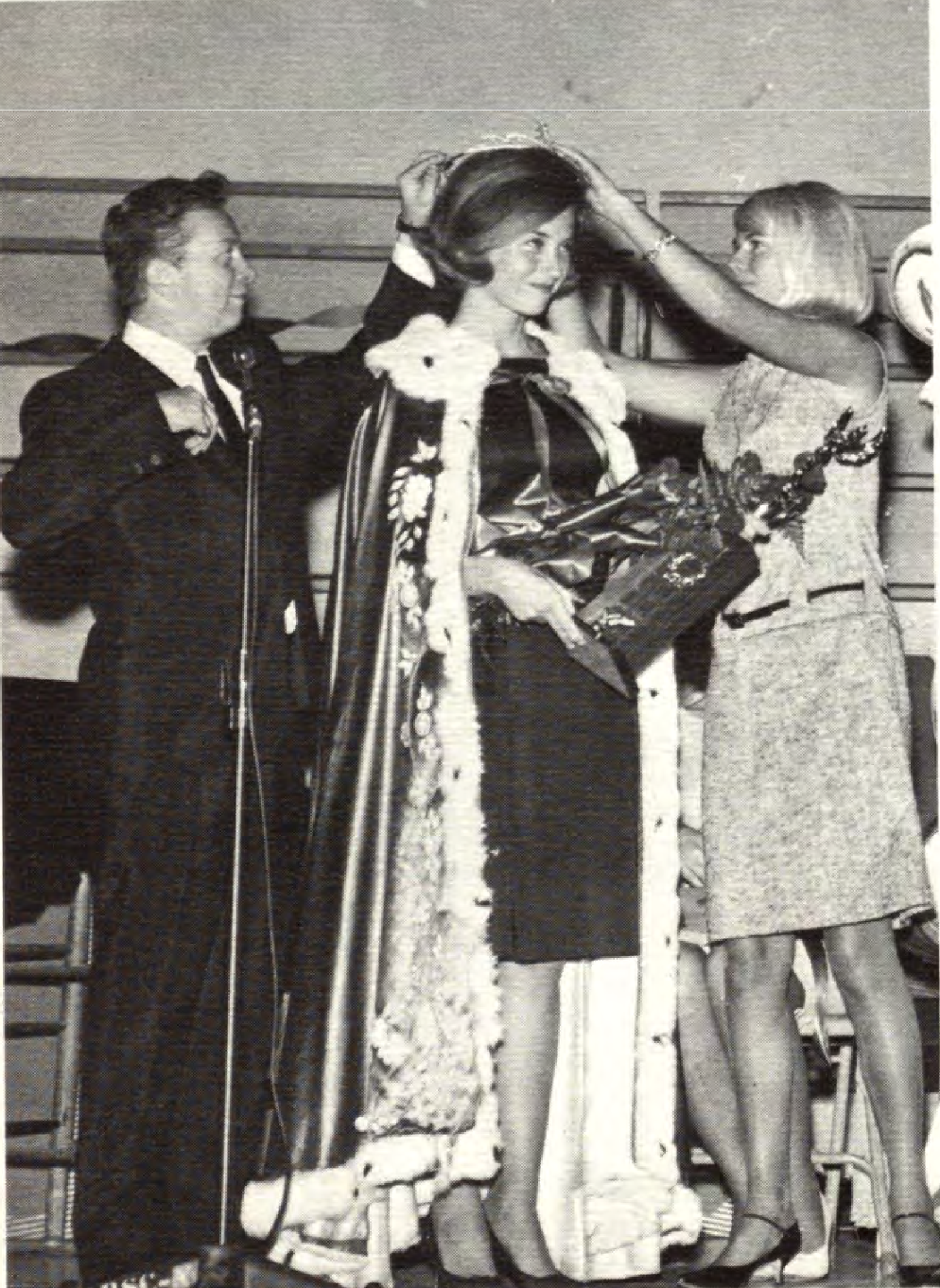 A person is crowned while standing in a fur-trimmed robe and holding a bouquet.