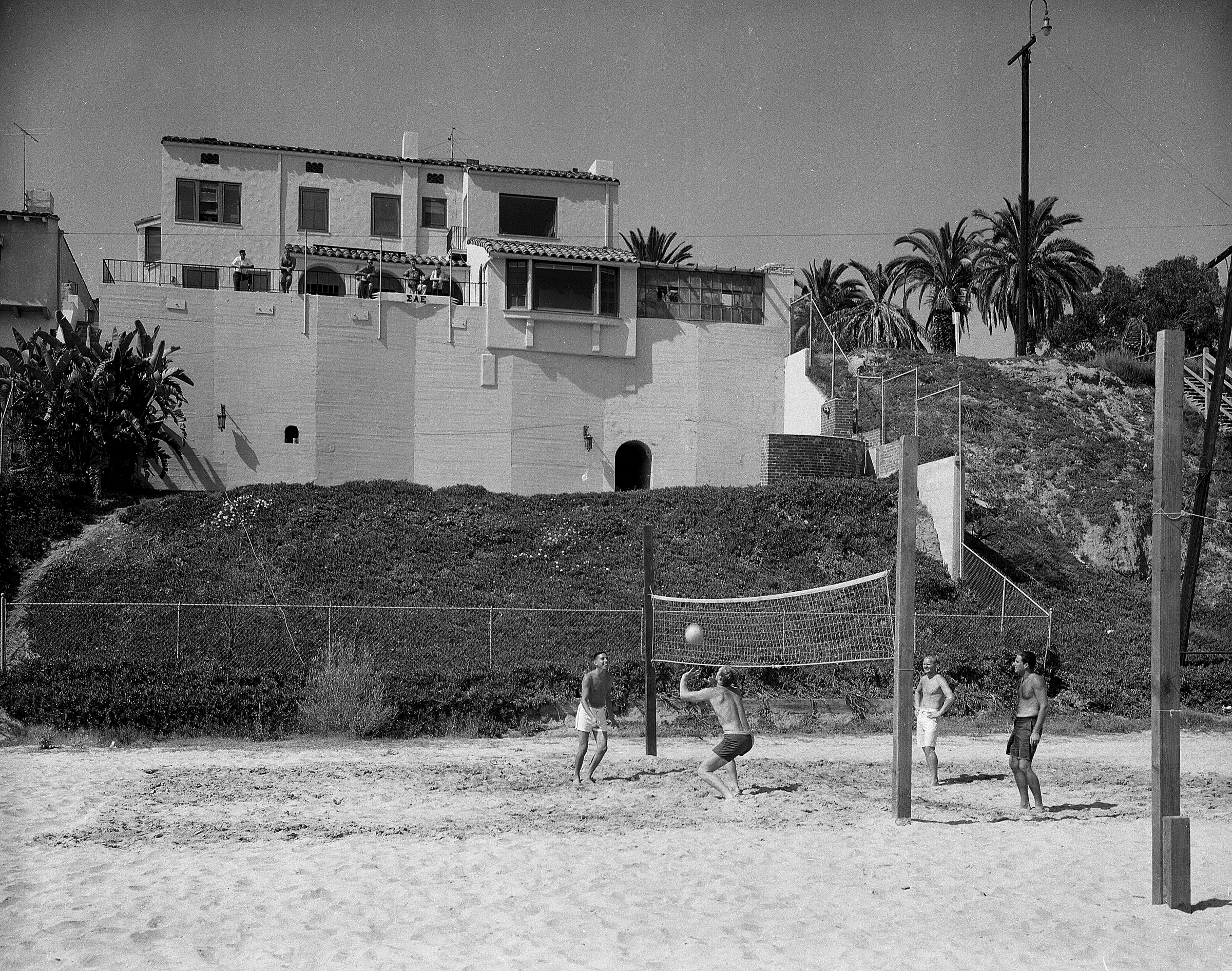 Four people play beach volleyball near a large house overlooking the beach.