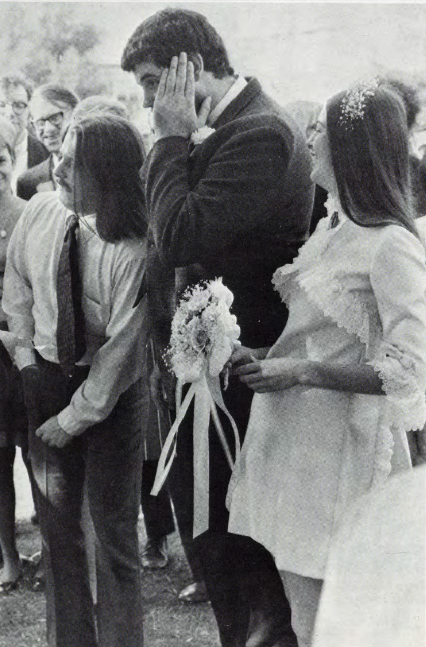 A newlywed couple and a guest stand together, the groom appearing distressed as another man rummages through his pocket.