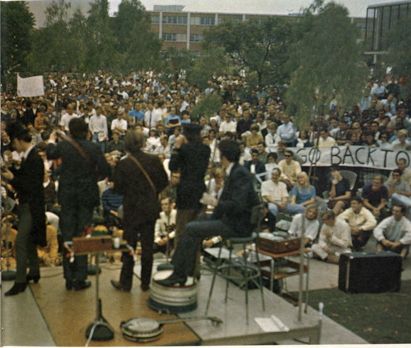 A band performs on a small stage in front of a large crowd outdoors.