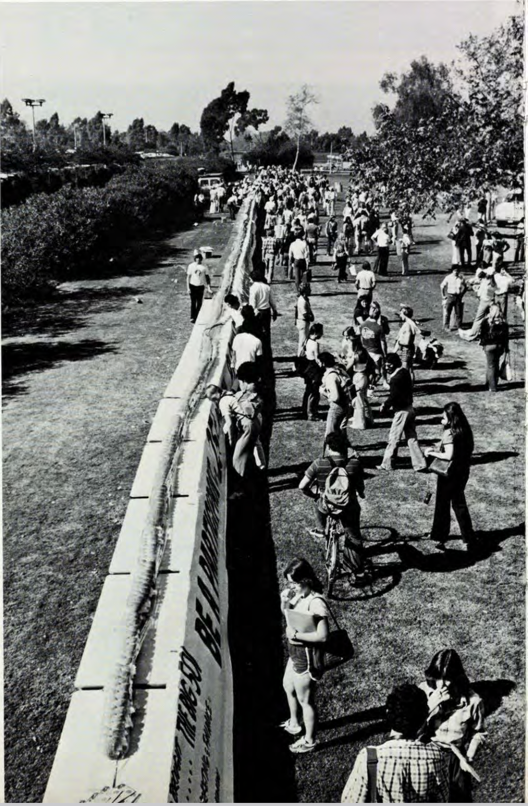 A crowd walks along a long a table with a long sandwich on it outside.