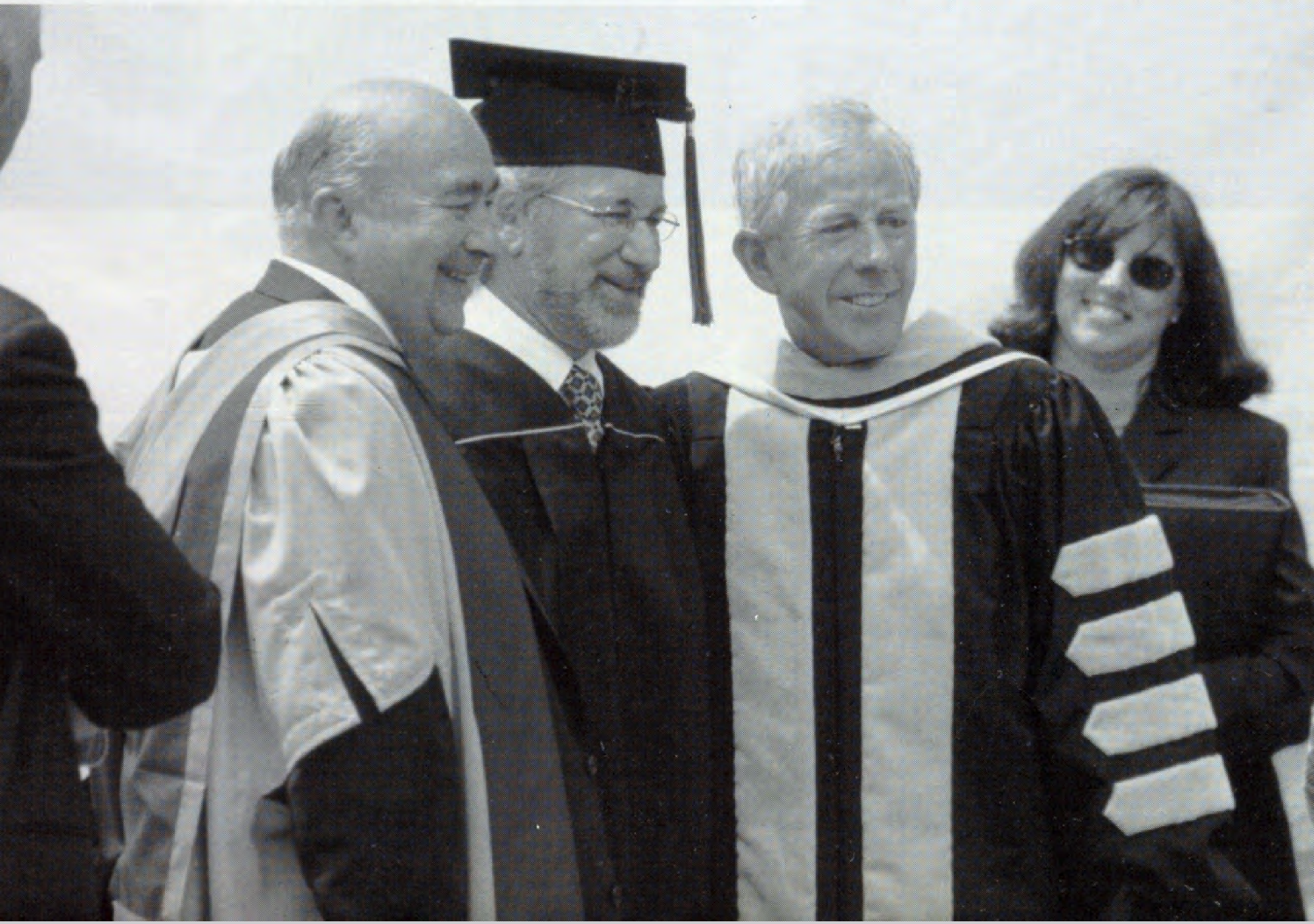 Three people in academic robes smile at a graduation ceremony.