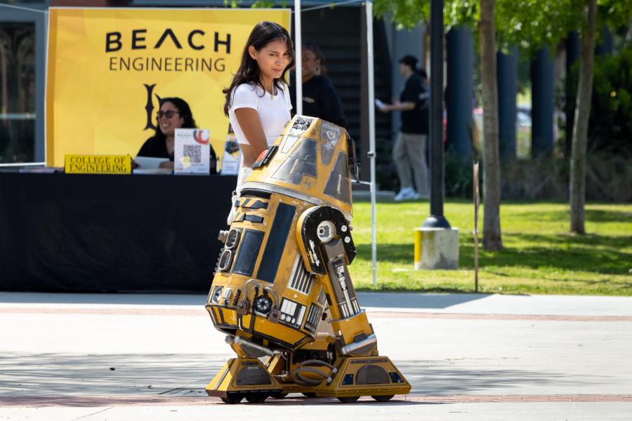 A student guides a small robot in front of a "BEACH Engineering" booth.