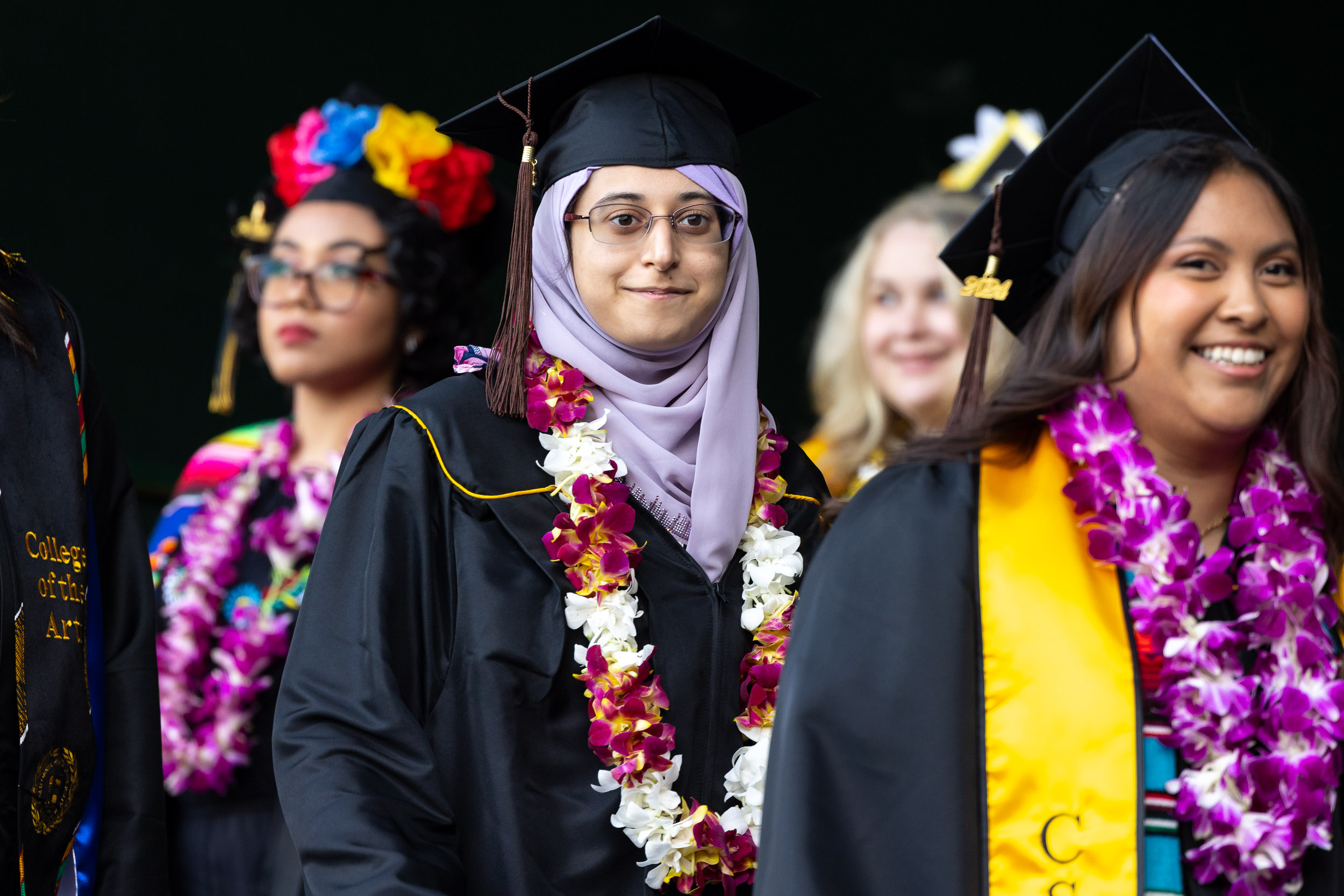 Graduates wearing caps and gowns stand in line, adorned with flower leis, smiling during a commencement ceremony.