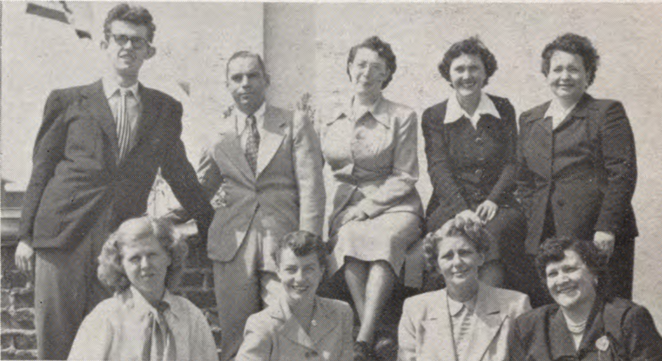 A group of eight adults in formal attire poses for a photo outdoors, with five standing and three seated.