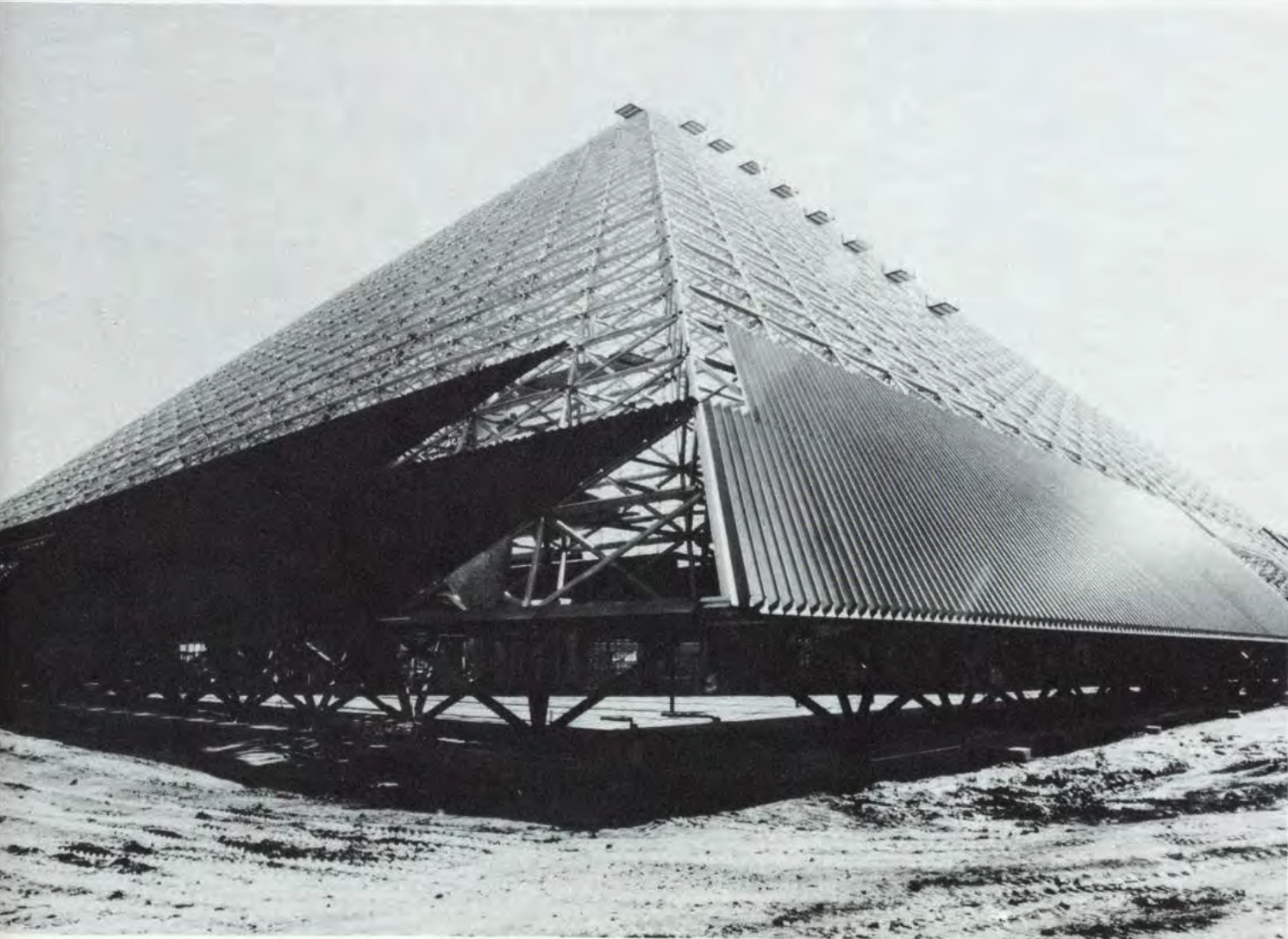 The steel frame of a large pyramid building under construction, set against a cloudy sky.