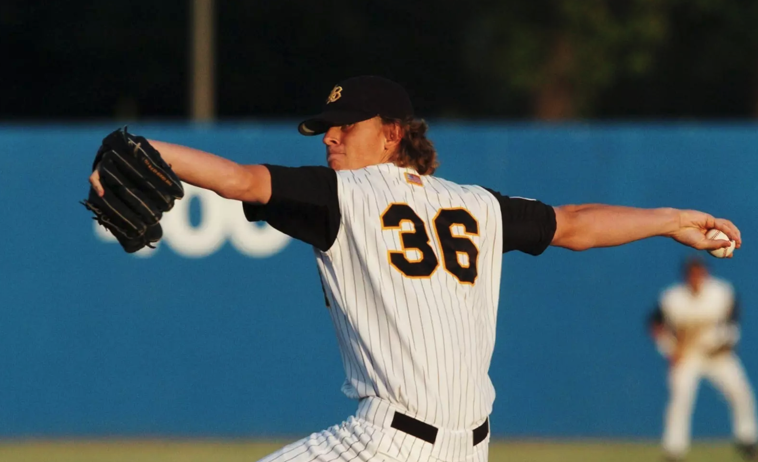 Jered Weaver in a white pinstripe uniform and black cap winds up to throw, with a fielder visible in the background against a blue wall.