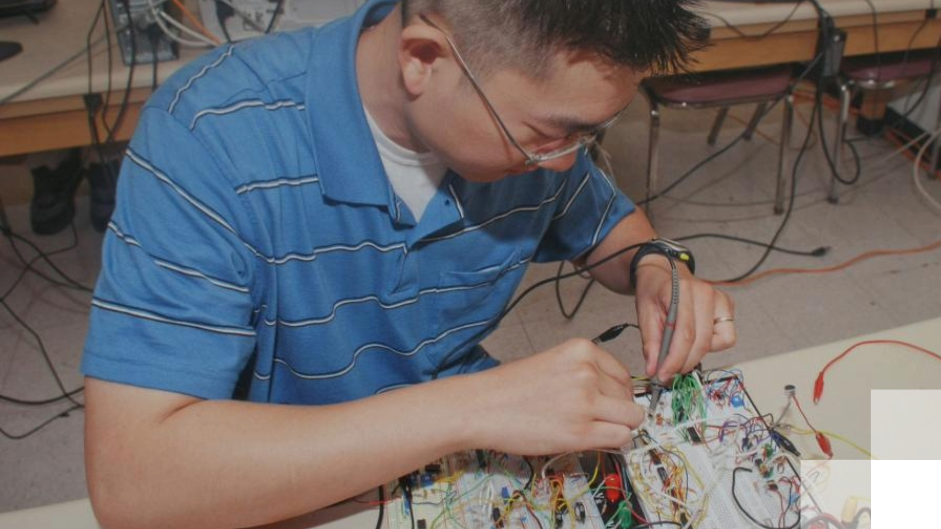 A man sits over a circuit board while holding wires. 