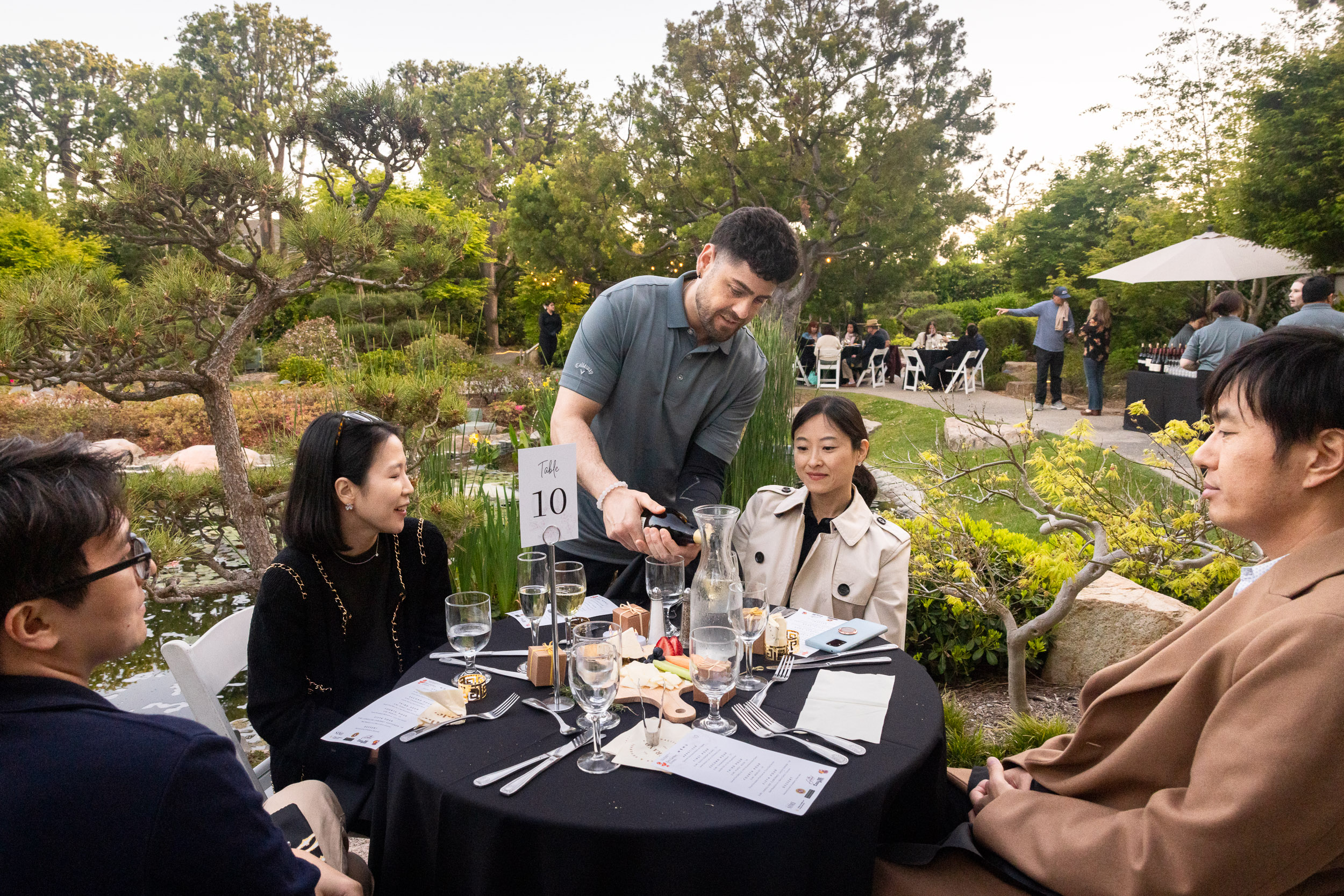 A server pours wine at a round table set with menus, glassware, and small plates, as guests sit and talk in a lush garden setting.