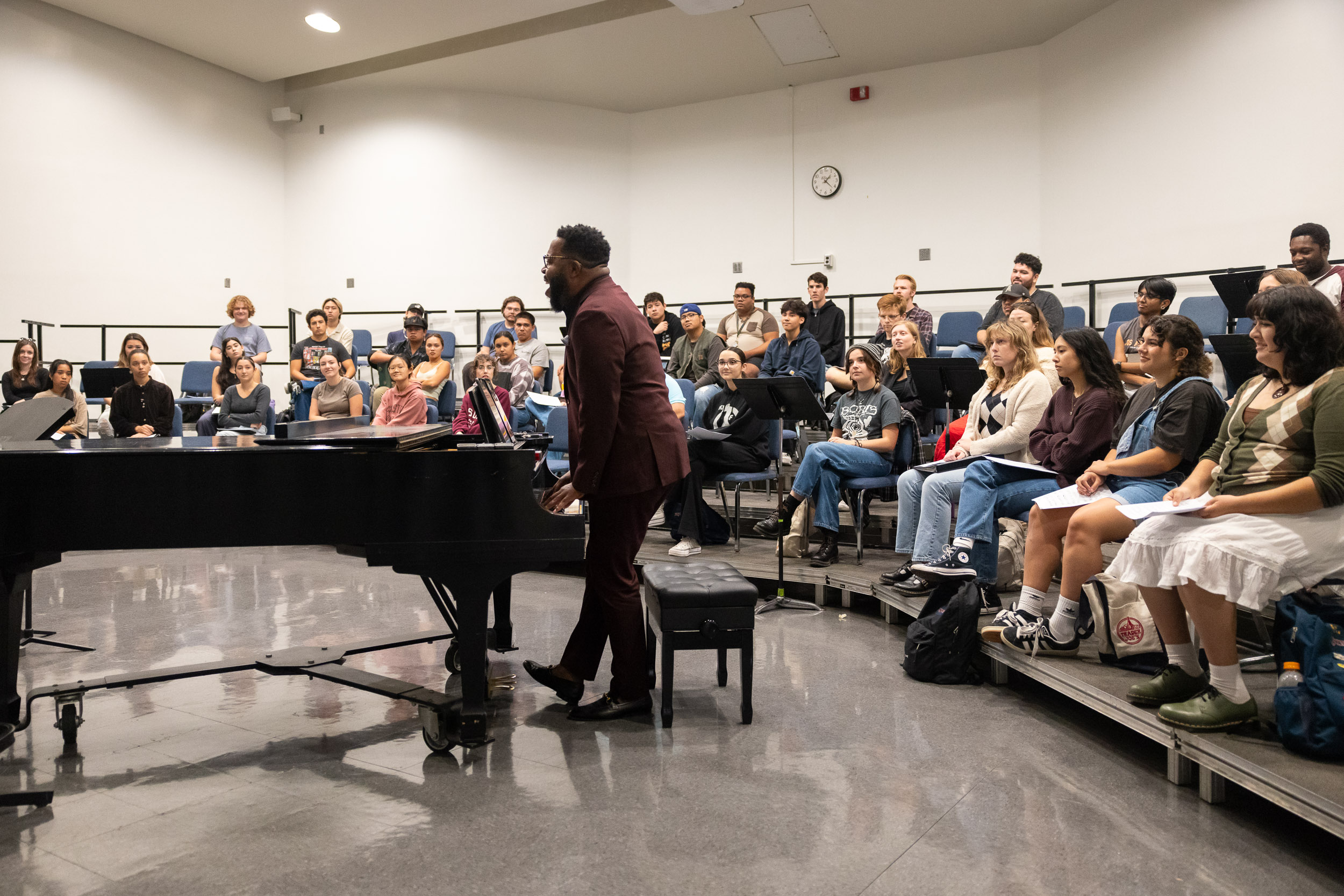 Musician Ananias Montague leads a rehearsal, standing and playing a piano, in a room full of seated students.