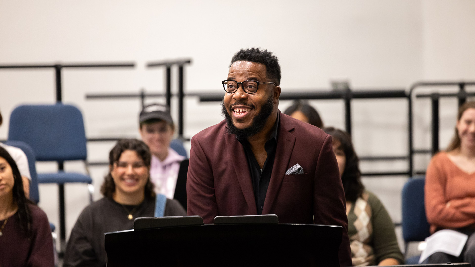 Ananias Montague, in a burgundy suit smiles while standing at a piano, surrounded by people in a rehearsal room.