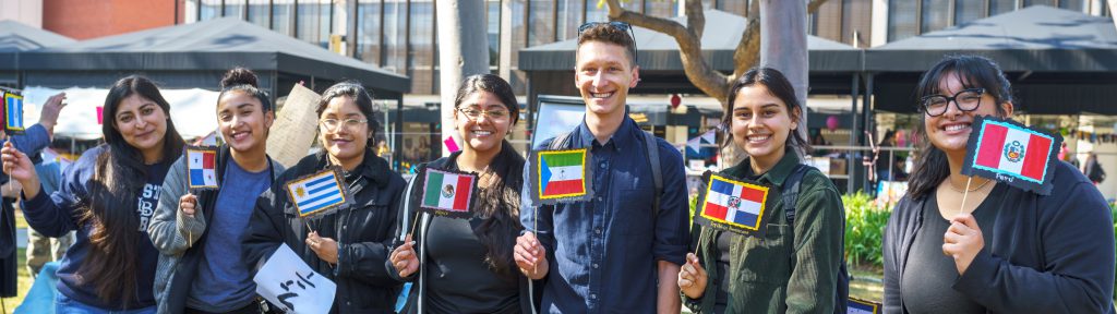 A group of students stand in a row facing forward, each holding a flag of a multicultrual background.