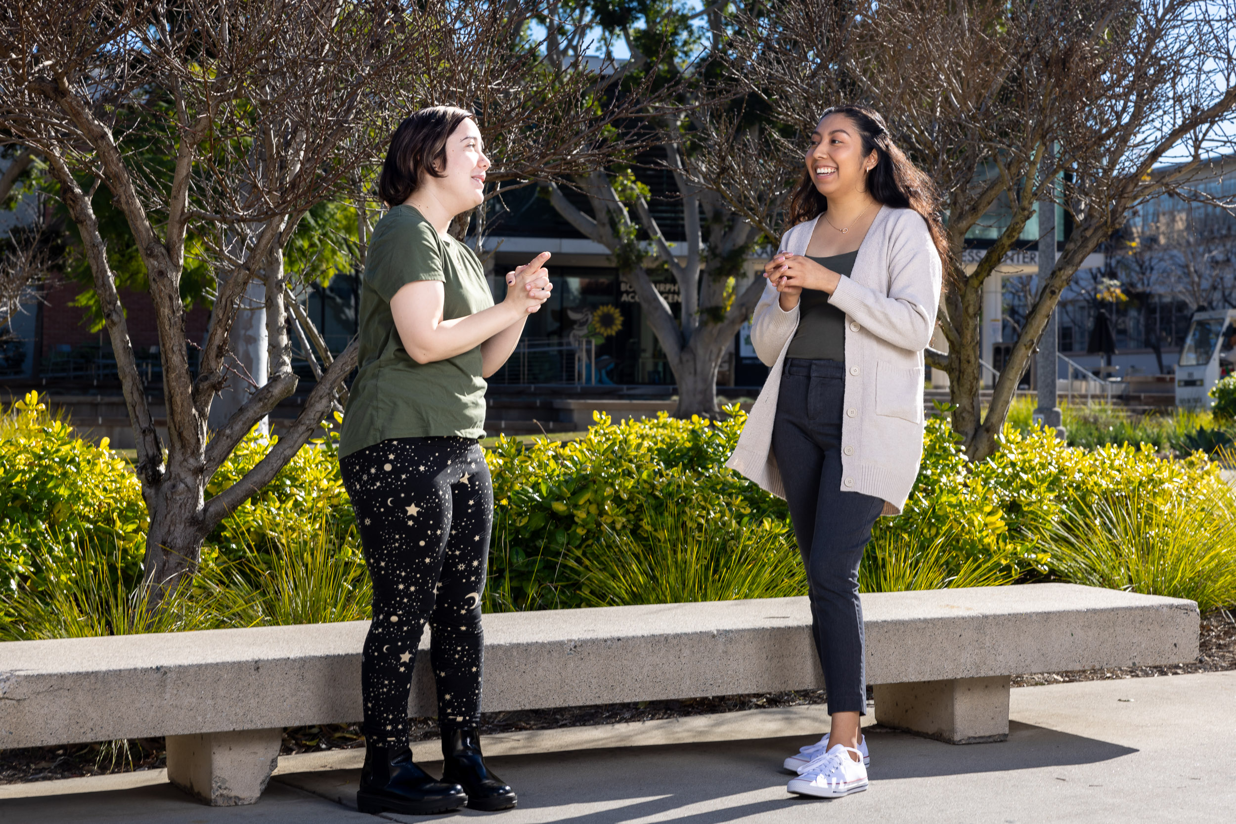 Teaching students Marisa Saxon and Yorgely Jimenez engage in conversation near a bench on campus