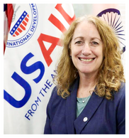 Karen Klimowski, a white woman with blond, shoulder-length hair and a blue blazer, stands next to a USAID flag