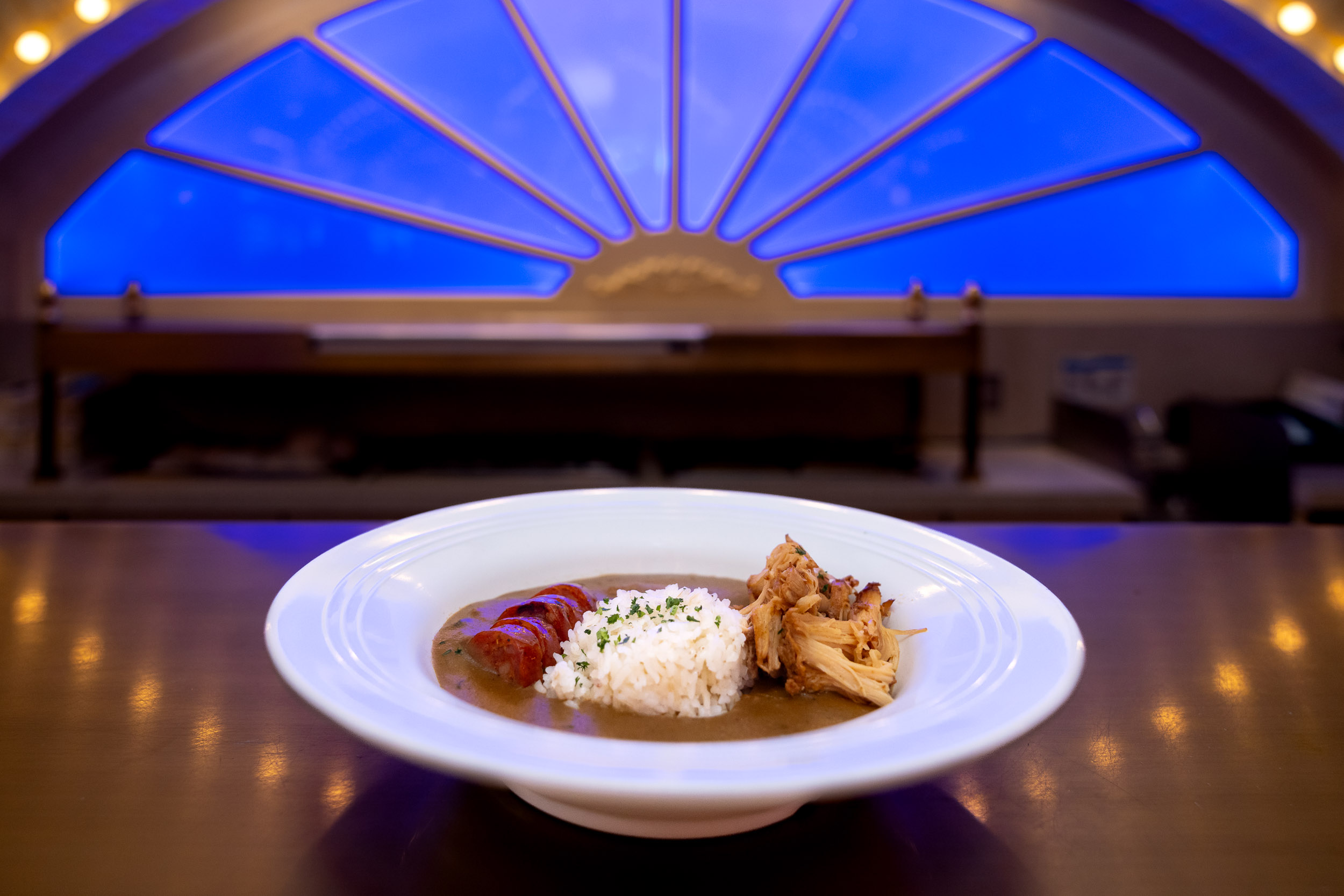 A plate of gumbo with rice, sausage, and shredded meat sits on a counter with a blue-lit window in the background.