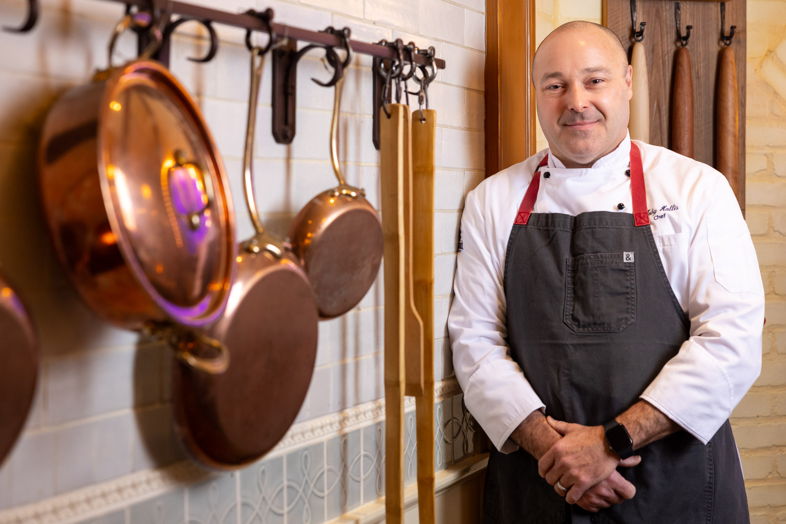 Toby Hollis stands beside a wall with copper pots and wooden rolling pins hanging