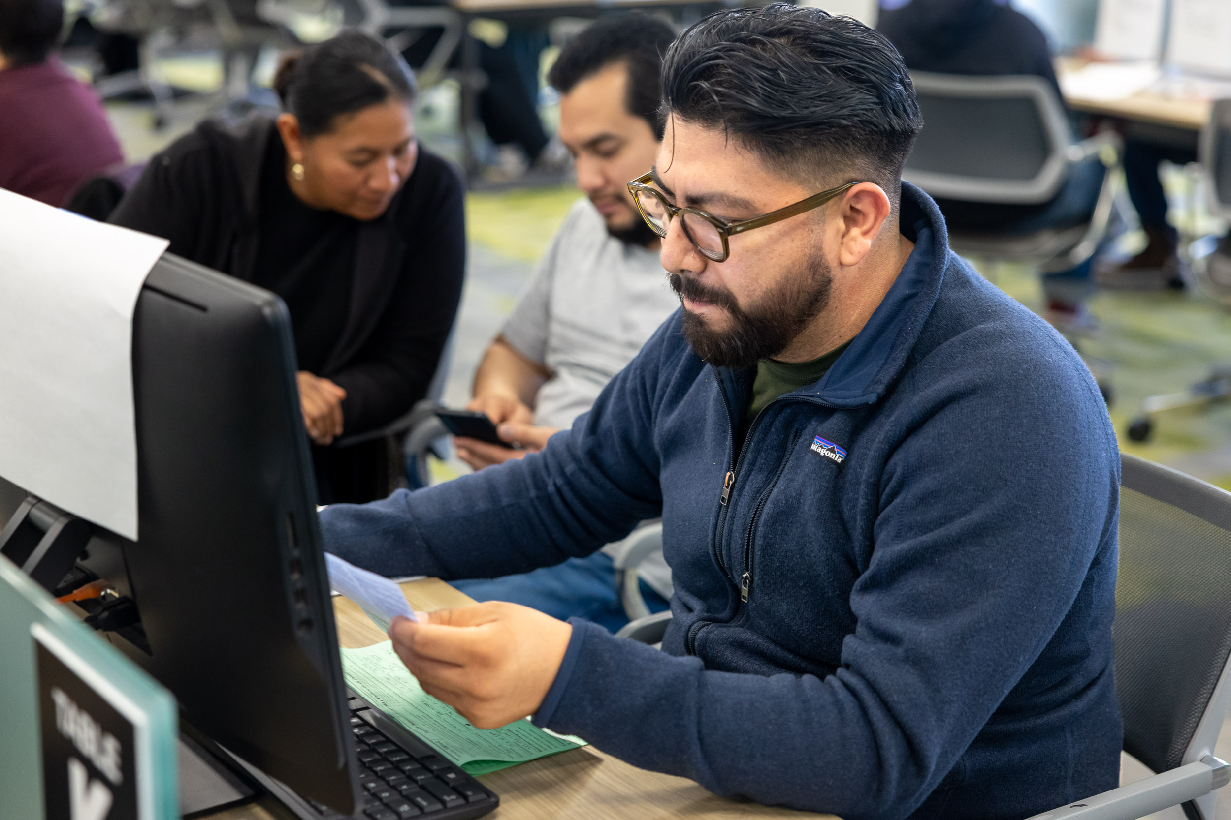 A bearded student in glasses examines a document while seated at a computer, with a couple behind him.