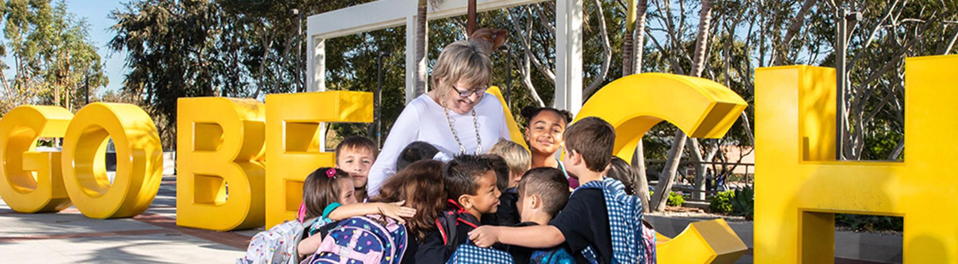 Conoley stands with children who are hugging her in front of "GO BEACH" sign, banner size Long Beach State University President Jane Close Conoley stands among a group of children with backpacks who are hugging her in front of large yellow "GO BEACH" letters on campus.