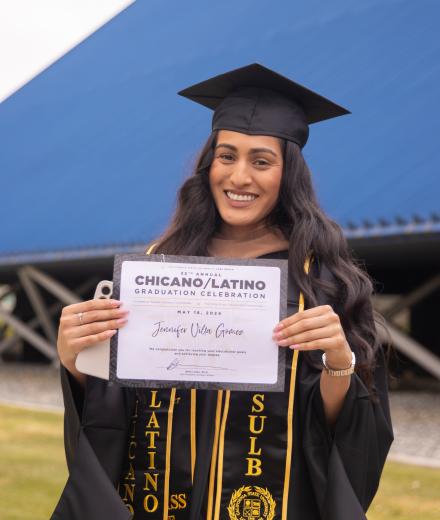 female graduate with a chicano/latino gradaute certificate and in her cap and gown. She is posing in front of the pyramid after the cultural graduation ceremony 