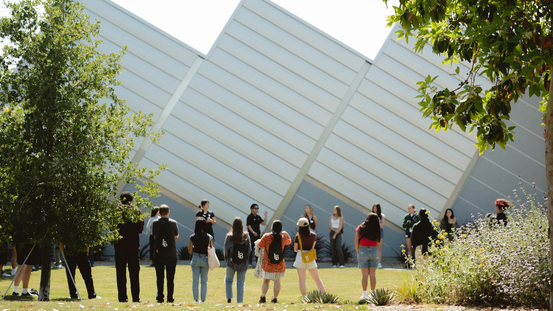 A group of people standing in a semi-circle outdoors, listening to a speaker in front of a modern, angular building.