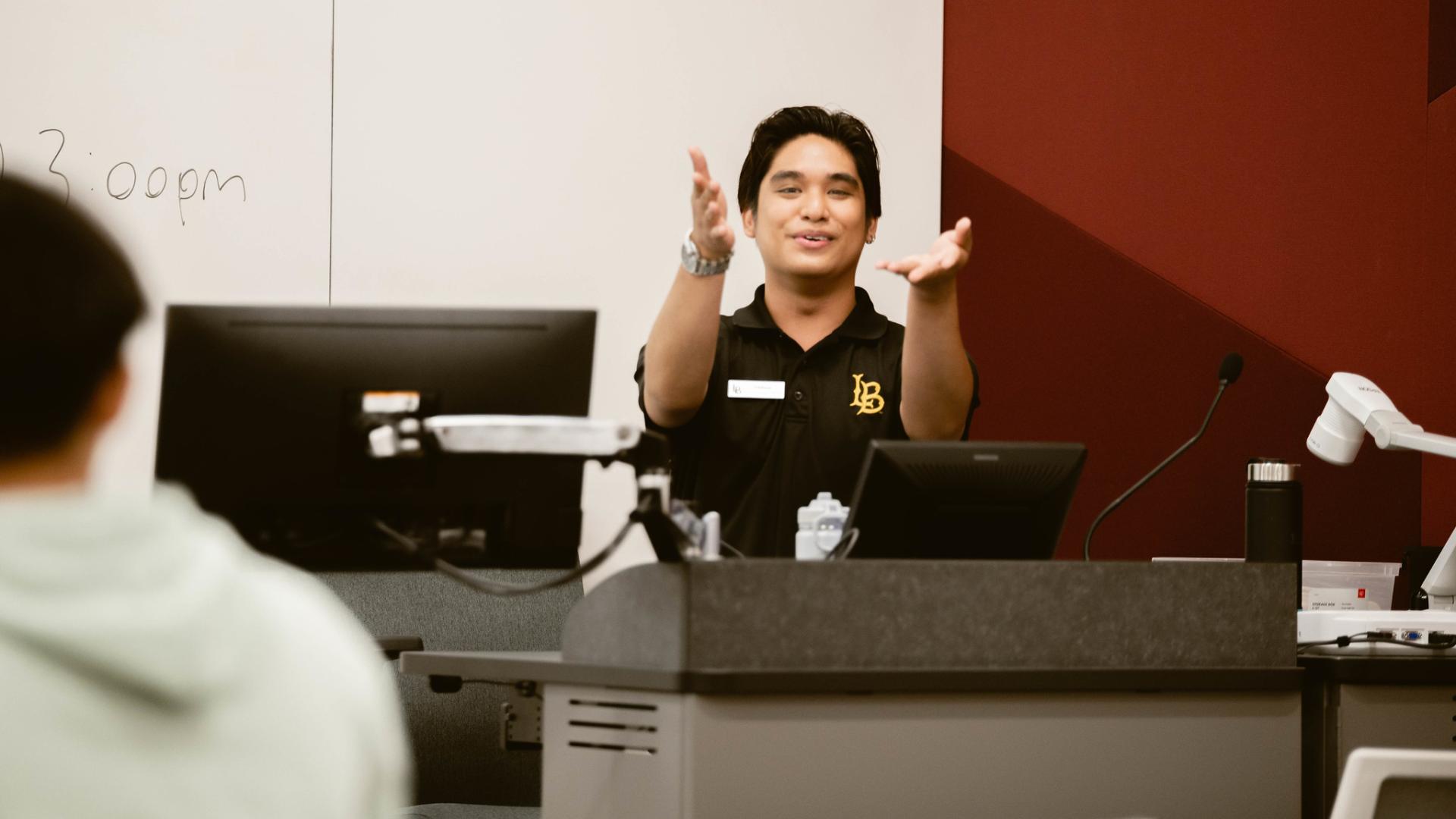A student leader standing at a podium in a classroom, gesturing with their hands while speaking to the audience.