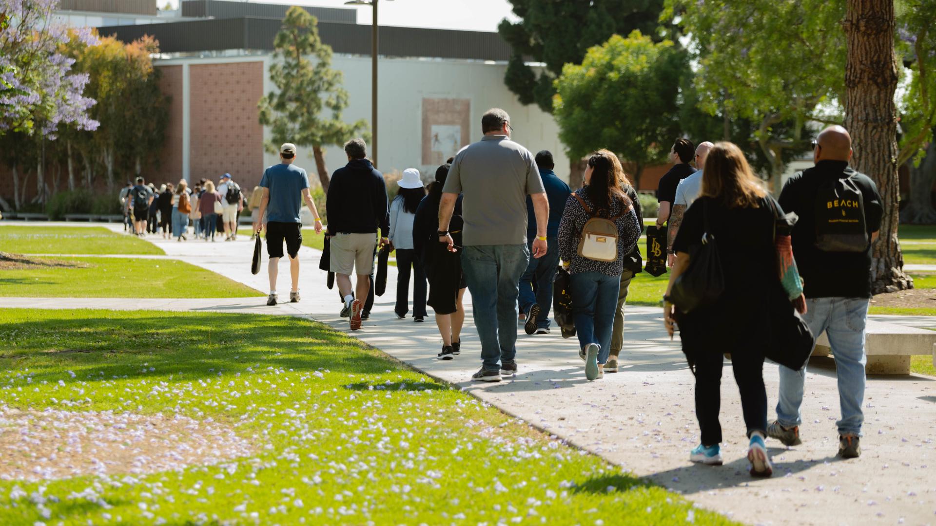 A group of people walking along a pathway on a green campus, heading towards a building in the background.