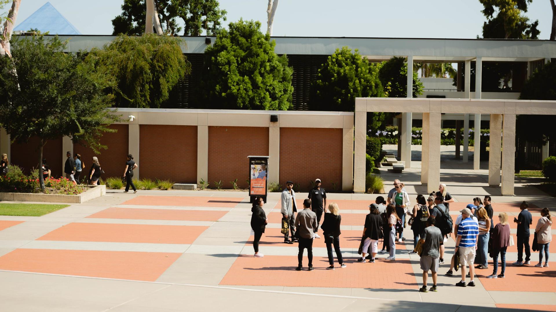 A group of people gathered in an outdoor courtyard, with some individuals walking in the background near a modern building with red brick and concrete columns.