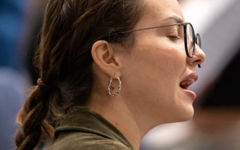 A student with glasses and a braid sings during a rehearsal.