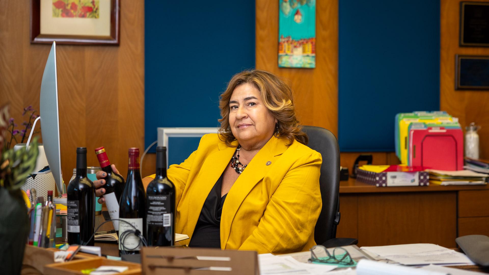 Anna Ortiz, wearing a yellow blazer, sits at a desk with a computer and several wine bottles, looking into the camera.