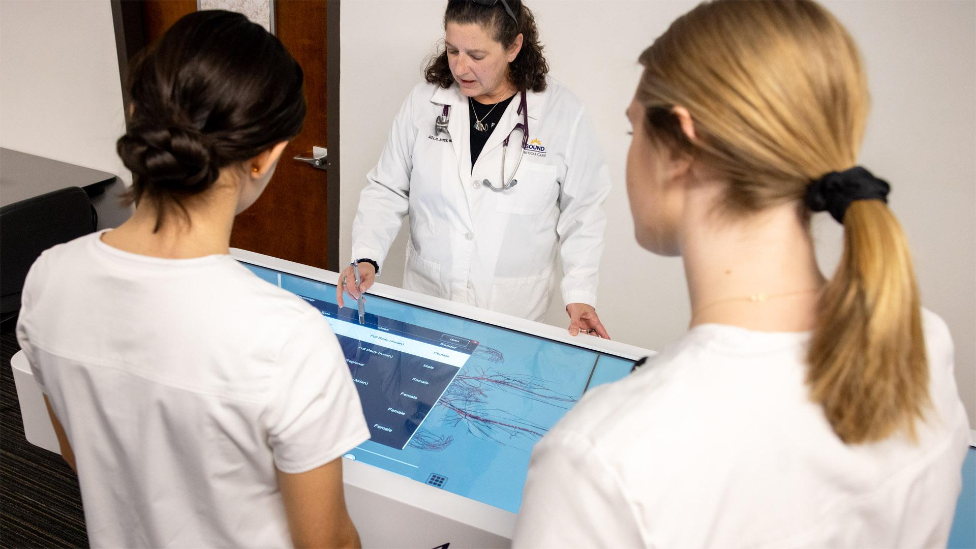 A School of Nursing faculty member in a lab coat demonstrates an interactive anatomy display to two students in white uniforms.