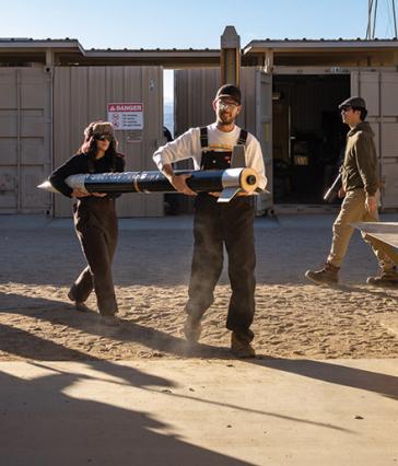 Two students carry a rocket body across a sandy outdoor workspace while others walk nearby.