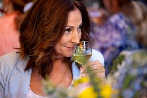  A person with shoulder-length brown hair smiles subtly while holding a glass of white wine at a gathering, with blurred people and flowers in the background.