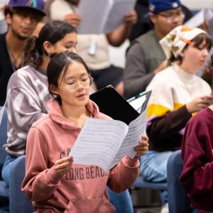 A student in a pink hoodie sings while holding sheet music, surrounded by others with music.