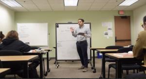 A man stands in front of a white board, teaching a class, with a marker in his hand.